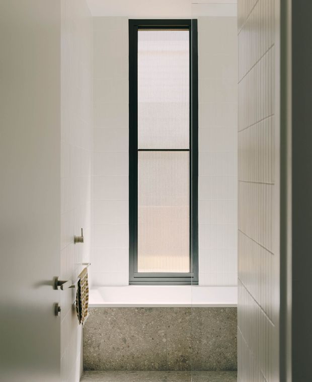 Compact modern bathroom with white tiled walls, a deep freestanding or built-in tub beneath a tall narrow frosted window, dark metal window framing, and a grey stone-look tiled bath surround. Minimal fittings and a neutral palette create a clean, understated space.