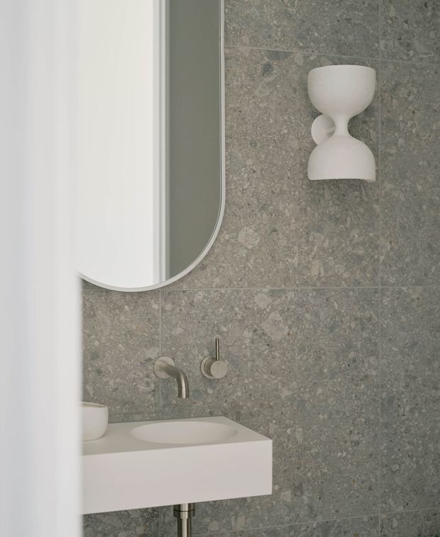 A refined contemporary bathroom vanity vignette viewed partly through a doorway or partition. The room features grey stone-look wall tiling, a white floating rectangular vanity with integrated basin, brushed metal wall-mounted tapware, a rounded-corner mirrored cabinet, and a sculptural white wall light. The palette is soft grey and white with minimalist detailing.