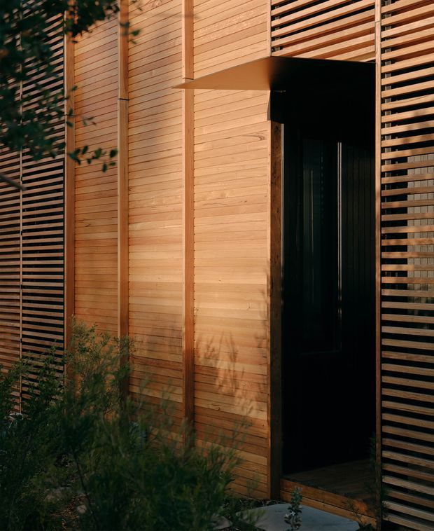 A close-up exterior view of a contemporary timber-clad entry facade. Warm horizontal wood boards and vertical framing surround a tall black doorway, with slatted timber screening on both sides and low planting at the base. The sheltered entry creates a strong contrast between the warm timber and dark door.