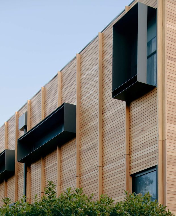 Exterior architectural photograph of a modern multi-storey building clad in pale horizontal timber boards, with projecting black metal box balconies and vertically expressed timber framing. A partial window is visible at the right, with low planting along the base and clear sky behind.