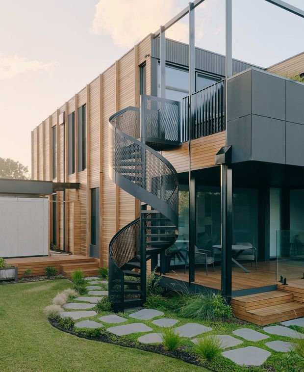 A contemporary multi-level residential exterior with warm horizontal timber cladding, dark metal framing, expansive glazing, and a prominent perforated metal spiral staircase connecting upper and lower terraces. A timber deck and glazed outdoor seating area open onto a landscaped garden with stepping-stone paving and grasses.