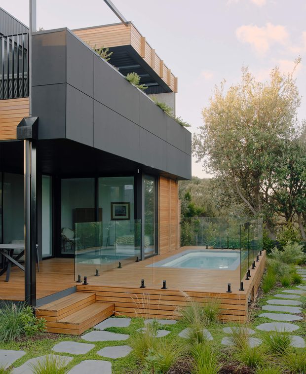 Contemporary multi-level residential exterior with dark panel cladding, warm timber decking and balcony screening, extensive glazing, and a rooftop terrace. The home opens onto a timber pool deck with frameless glass balustrades, surrounded by landscaped planting and stepping stones.