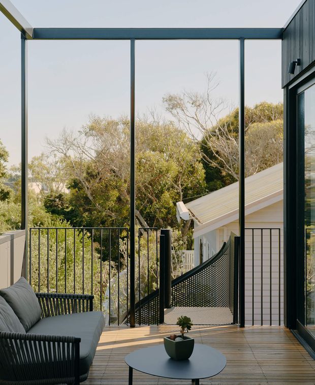 A contemporary outdoor balcony or terrace viewed from beside a glazed building facade. The space features a timber deck, slender black metal balustrades, full-height dark-framed glazing, a dark woven outdoor sofa with cushions, and a small round coffee table with a potted plant. Trees and a neighbouring sloped roof are visible beyond the railing, creating a leafy elevated outlook.