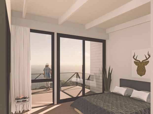 A bright, contemporary bedroom with a dark quilted bedspread, white pillows, a black headboard, and minimalist deer artwork. Full-height black-framed glazed doors open onto a balcony or terrace, with sheer curtains, timber decking, and a hazy exterior view. White walls and exposed ceiling beams create an airy, refined interior.