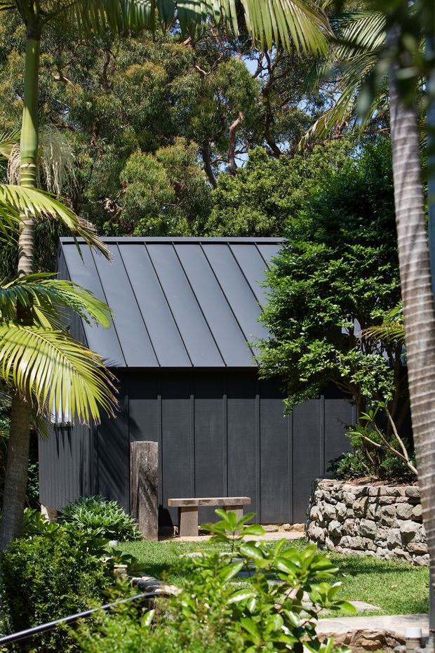 A compact dark-clad garden outbuilding or pavilion is set within dense tropical landscaping. The structure has a steeply sloping standing-seam metal roof, charcoal vertical wall cladding, and a weathered timber door. A simple timber bench, stone retaining wall, lawn, and stepping stones occupy the foreground, framed by palms and lush subtropical vegetation.