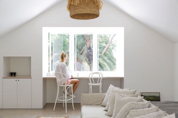 Bright, minimalist bedroom in a small gabled space, featuring white wall and ceiling panelling, a large central window overlooking greenery, and a built-in window-side desk or counter with two white stools. A white bed with layered neutral bedding occupies the foreground, while a woven pendant light adds natural texture.