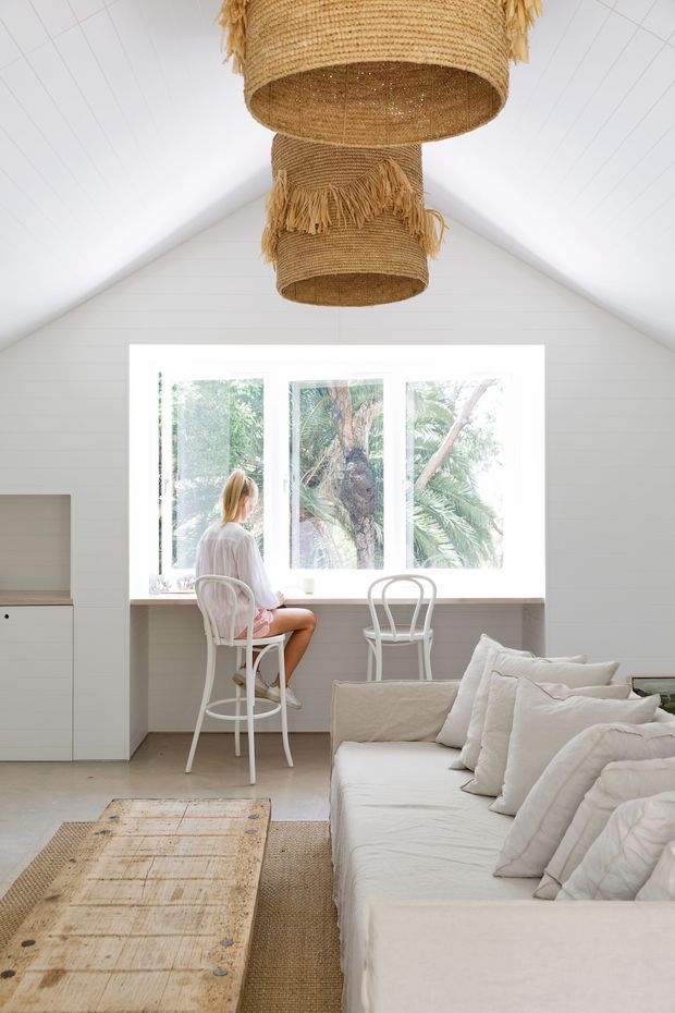 Bright, airy open-plan living space with a white-painted pitched ceiling and walls, large picture windows overlooking lush greenery, a white built-in window bench with two bar stools, and a white upholstered sofa in the foreground. Natural woven pendant lights, a jute rug, and a rustic timber coffee table add warm texture to the otherwise light neutral interior.