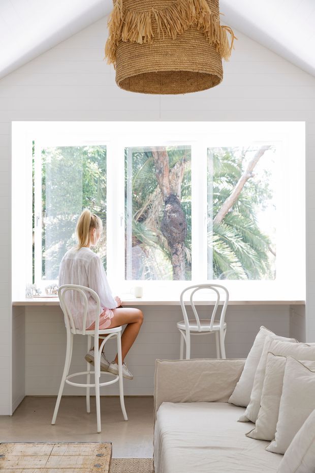 Bright, airy interior with white horizontal wall panelling, a pitched ceiling, and large white-framed windows overlooking lush vegetation. A woven natural-fibre pendant hangs above a compact built-in window bench or desk, accompanied by white bentwood-style chairs. A white upholstered sofa with layered cushions occupies the foreground, creating a relaxed coastal living space.
