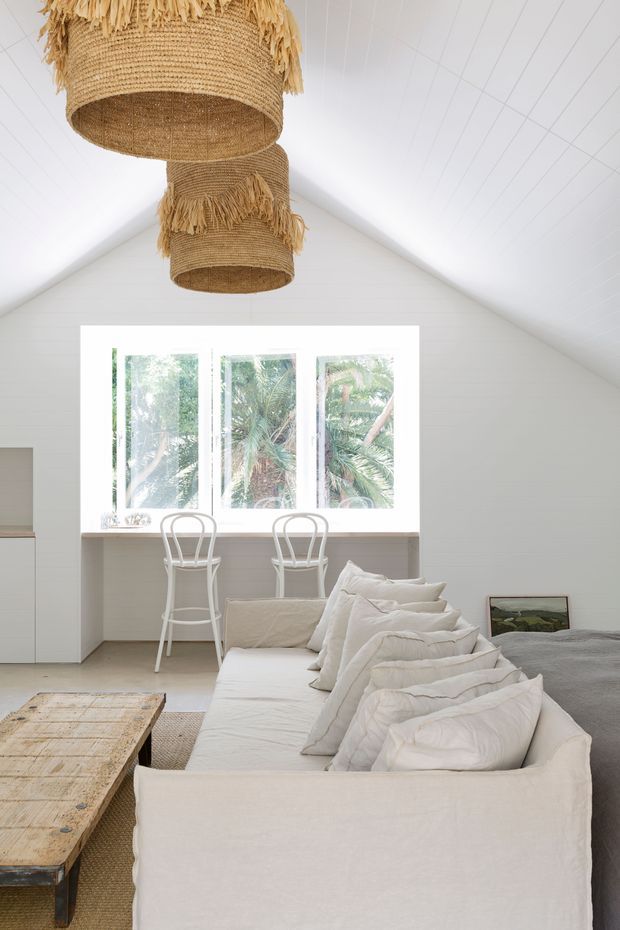 Bright, airy open-plan room with a white pitched, timber-lined ceiling and walls. Two large woven natural-fibre pendant lights hang above a white sofa, while a long built-in window seat or desk with two white bar stools sits beneath a large window overlooking trees. A rustic timber coffee table and soft neutral furnishings complete the relaxed interior.