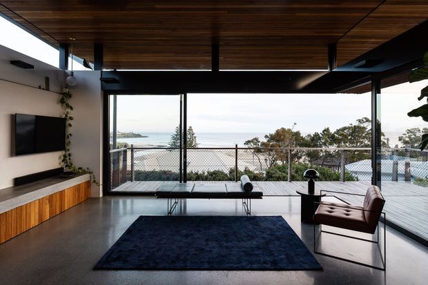 Modern open-plan living room with full-height glazing overlooking a coastal or waterfront setting. The room features a dark timber slatted ceiling, polished concrete floor, built-in timber media cabinetry, black-framed sliding glass doors, a dark rug, a low coffee table, and a leather lounge chair. An expansive timber deck and outdoor railing extend beyond the glazing.