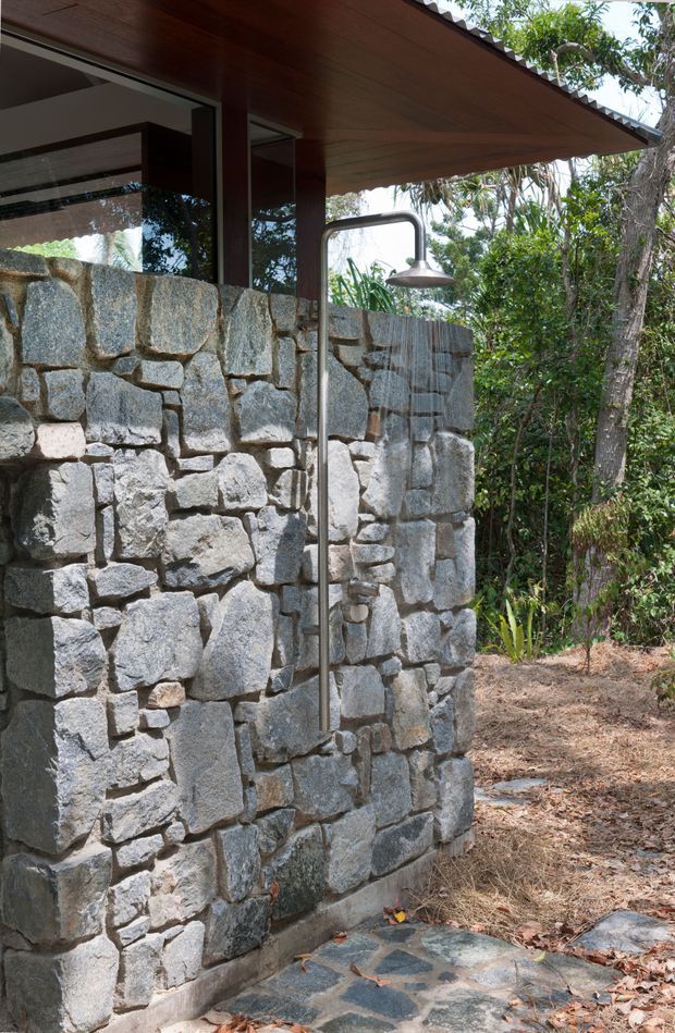 Exterior view of a rustic outdoor shower beside a substantial rough-stone masonry wall. A dark metal shower pipe and round showerhead stand beneath a broad timber-lined roof overhang, with large glazed openings visible behind the wall. The setting is surrounded by dense natural vegetation and a stone-paved threshold, creating an earthy, integrated indoor-outdoor bathing area.