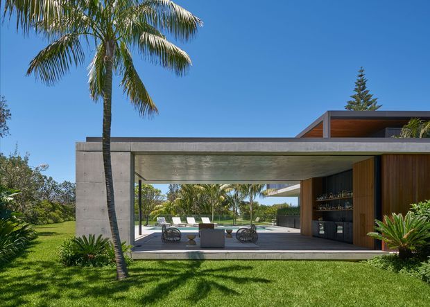 A contemporary single-storey pavilion-style home with a strong indoor-outdoor connection. The image shows a broad exposed concrete frame enclosing a shaded outdoor living area, with timber-clad walls and a built-in bar or joinery zone on the right. A flat roof and deep overhang extend over a paved terrace furnished with outdoor seating, while landscaped lawn and tropical planting surround the structure.
