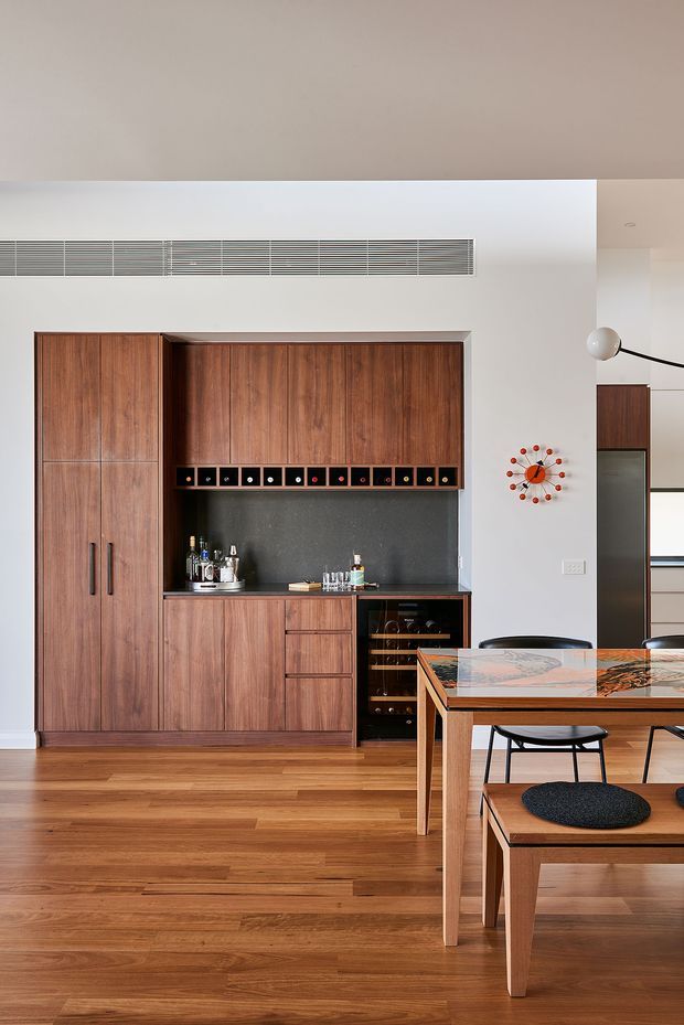 A warm, refined dining area with a built-in walnut-toned cabinetry and bar wall, including tall storage cupboards, open shelving for bottles, a dark stone-look backsplash and benchtop, and an integrated wine refrigerator. A timber dining table with a richly patterned stone or marble-look top and matching timber chairs occupies the foreground. White walls and ceiling, timber flooring, a linear air vent, a mid-century-style wall clock, and a sculptural floor lamp complete the interior.