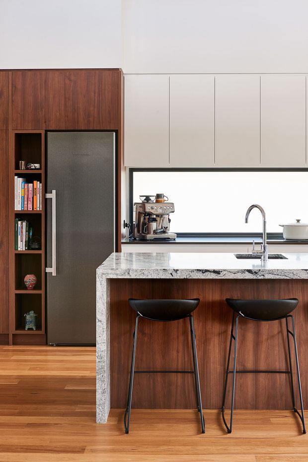 A refined contemporary kitchen featuring dark timber cabinetry and integrated open shelving, a stainless-steel refrigerator, pale upper cabinets, and a large island with a heavily patterned stone benchtop. Two black bar stools sit at the island, while a coffee machine, sink, and cookware are visible along the rear counter. Warm timber flooring and cabinetry contrast with the light walls and cabinetry.