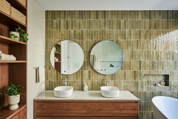 A contemporary bathroom featuring a wide timber vanity with two white vessel basins, round mirrors, and brushed metal tapware. The walls are clad in vertically arranged glossy olive-grey ceramic tiles, while open timber shelving with woven baskets and plants occupies the left side. A freestanding white bathtub is partially visible at right, creating a warm, natural and refined atmosphere.