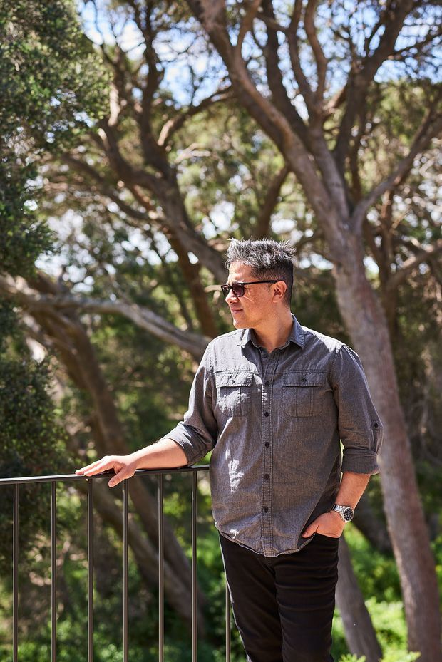 Portrait of a man standing on a raised outdoor balcony or terrace, one hand resting on a simple dark metal balustrade. The setting is surrounded by mature trees and dense greenery, with bright natural daylight and a softly blurred woodland background. The image is primarily a lifestyle portrait rather than an architectural or interior space photograph.