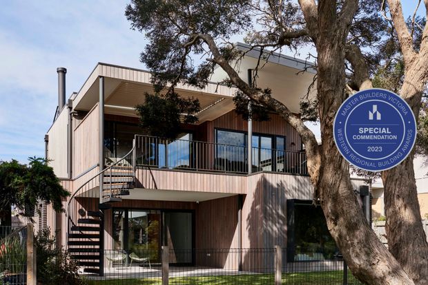 Exterior view of a contemporary multi-level residence with vertical timber-like cladding, broad roof overhangs, extensive glazing, and a prominent external spiral staircase leading to an upper balcony. Mature trees partially screen the facade, and a builders-award sign is visible in the foreground.
