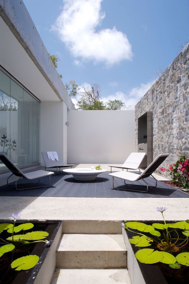 A bright, minimalist outdoor courtyard terrace beside a shallow rectangular water feature with lily pads. The terrace has a dark textured surface, white rendered walls, a large frameless glass opening, four low modern lounge chairs, and a sculptural white coffee table. A rugged exposed stone wall with a recessed doorway forms one side, while planted edges and a clear blue sky create an airy contemporary setting.
