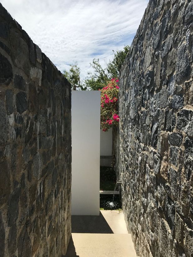 A narrow outdoor passage between two tall, rugged dark-stone masonry walls. A minimalist smooth white vertical wall or gate stands at the end, with a vivid flowering shrub visible behind it. Concrete or pale paved steps lead through the shaded, textural corridor into a small landscaped area. The image has a strong contrast between rough natural stone and clean modern geometry.