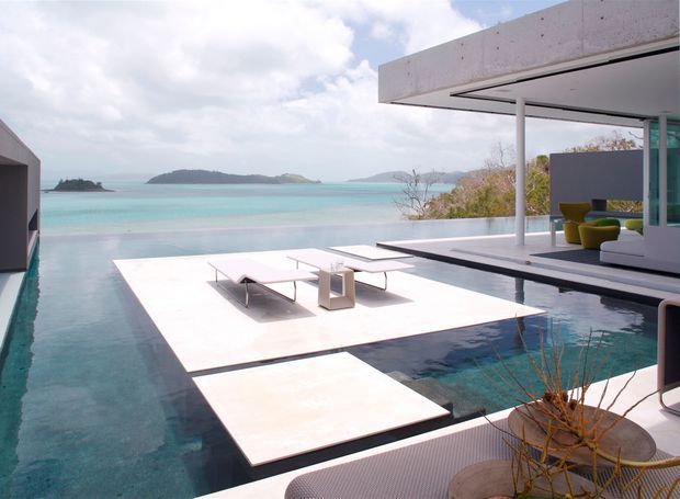 A minimalist modern poolside terrace overlooking a turquoise bay with small islands. Large pale stone or concrete floating platforms extend over the pool and hold low-profile outdoor loungers and a small side table. The open-plan pavilion has a flat overhanging roof, slender white structural columns, expansive glazing, and a shaded lounge area with bright yellow seating. The scene is bright, serene, and strongly connected to the coastal landscape.