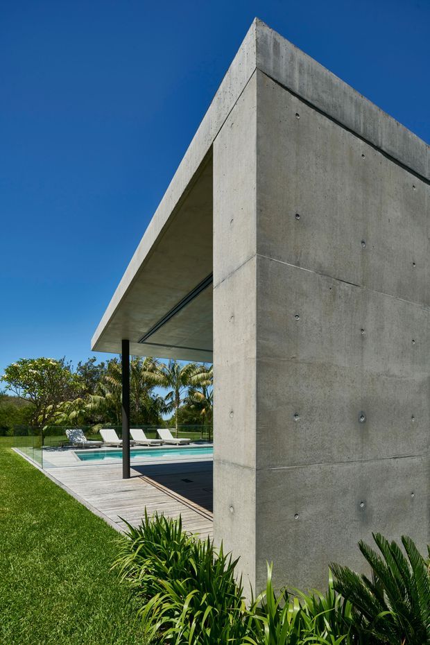 Exterior architectural photograph of a minimalist modern concrete pavilion beside a swimming pool. The composition highlights a tall board-marked concrete wall and projecting flat-roof canopy supported by a slender dark column, with timber pool decking, glazing, lounge chairs, tropical planting and bright blue sky visible around it.