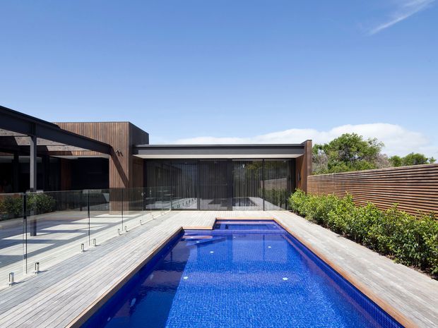 A contemporary residential courtyard viewed from the pool, featuring a long rectangular blue swimming pool surrounded by pale timber decking, frameless glass balustrades, dark metal roof structures, timber-clad walls, extensive glazing, and horizontal timber screening with planted borders. The scene is bright and open under a clear blue sky.