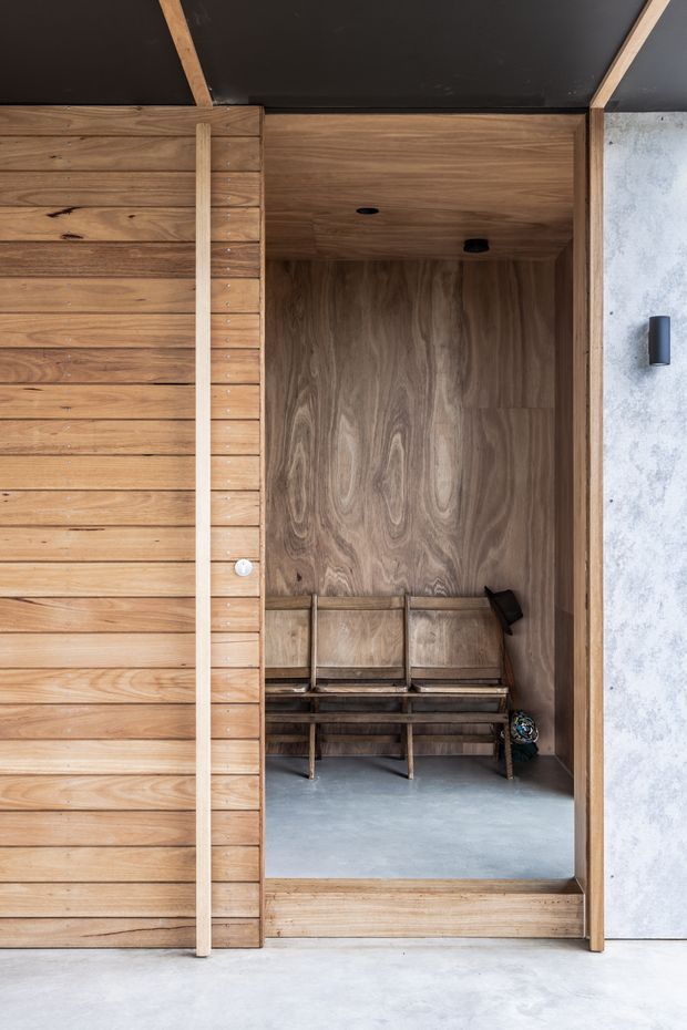 A compact timber-lined sauna room viewed from the entrance. Vertical and horizontal wood panelling surrounds a built-in wooden bench, with a dark ceiling, recessed lighting, and a smooth grey concrete or stone floor. The exterior edge combines timber cladding with a pale textured wall finish, creating a warm minimalist architectural interior.