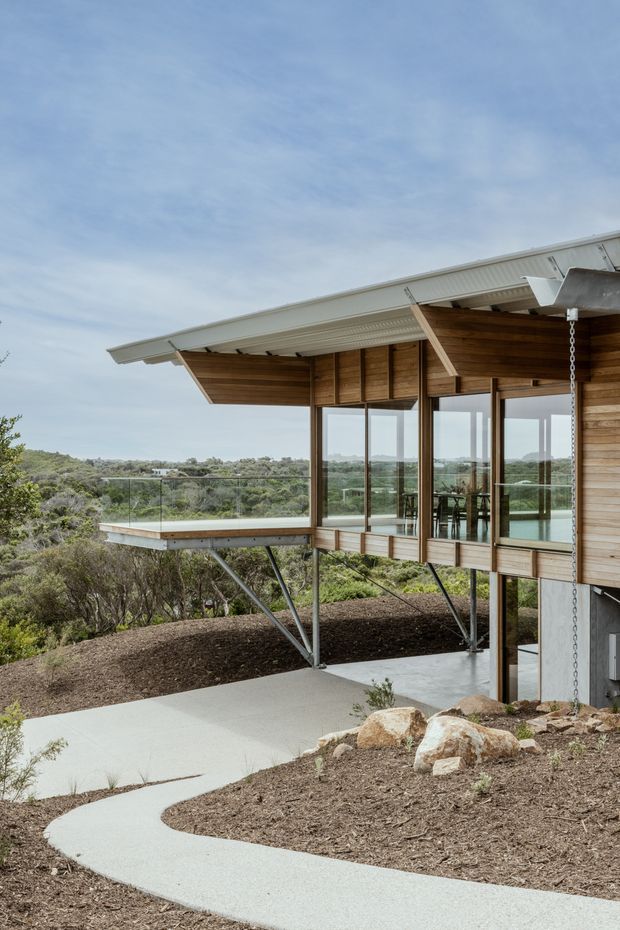 Exterior architectural photograph of a contemporary elevated timber-and-glass building set among native bush on a sloped site. The structure features a projecting balcony with clear glass balustrades, exposed steel supports, extensive glazing, horizontal timber cladding, and a broad low-pitched metal roof with deep overhangs. A curved concrete walkway approaches the building through the landscaped terrain.