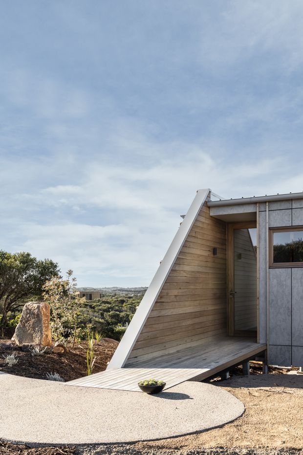 Exterior architectural photograph of a compact contemporary building set in a landscaped, semi-rural site. The structure features a steep triangular gable-like entrance form with pale timber horizontal cladding, grey fibre-cement or concrete-look wall panels, a timber-framed window, and a timber deck ramp leading to the recessed entry. Native-style planting, exposed rock, and a distant rolling landscape surround the building under an overcast sky.