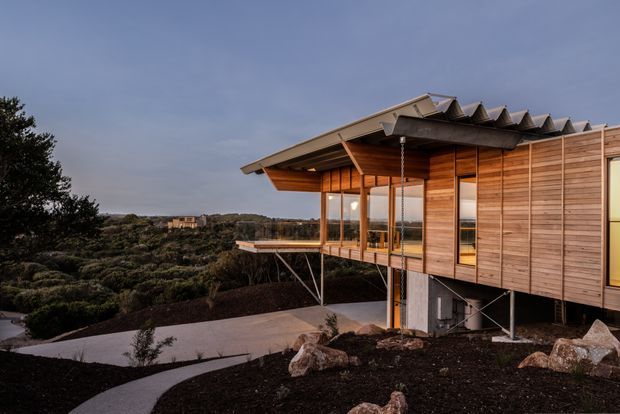 Exterior view of a contemporary elevated timber-clad house set on a landscaped sloping site. The building features extensive glazing, a projecting covered deck or veranda, slender support columns, a broad low-pitched roof with a corrugated-looking edge detail, and exposed structural elements. Curved concrete pathways and large rocks lead through the dark landscaped grounds, with a wooded landscape beyond.