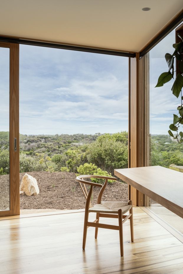 A bright, refined corner dining or work area with full-height timber-framed glazing overlooking a natural landscaped setting. Pale timber flooring, a simple light-toned built-in or cantilevered table, and a sculptural timber chair create a minimal connection to the outdoors. A leafy indoor plant is visible at the right edge, while the broad glazing provides expansive landscape views.