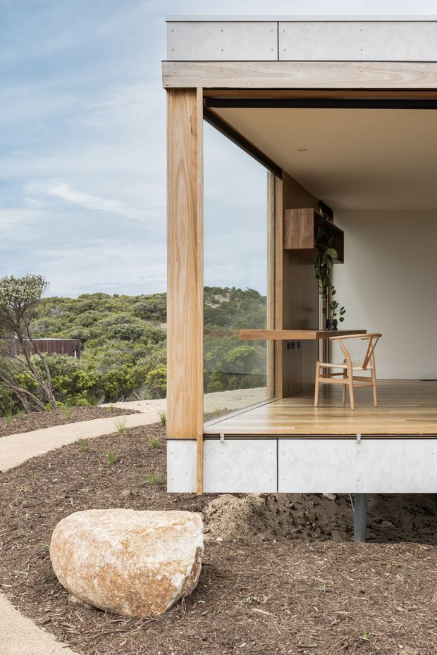 A modern elevated pavilion or small dwelling set within a natural bush landscape. The open corner reveals a compact interior workspace with a timber desk, light timber chair, built-in timber shelving and a trailing plant. Full-height glazing, warm timber framing and flooring, pale concrete-panel cladding, and a flat roof create a refined indoor–outdoor connection. A planted path and large natural boulder occupy the foreground.