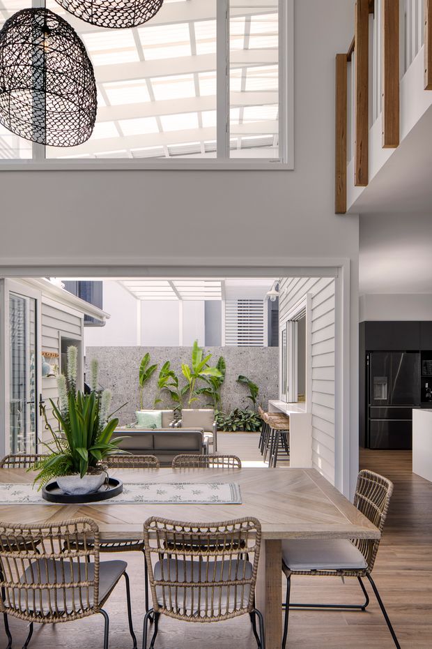 A bright open-plan dining area overlooking a planted courtyard through a wide opening. The room features a large light timber dining table with woven rattan dining chairs, pendant lights with black woven shades, white weatherboard-style walls, and large internal windows bringing in abundant daylight. A compact kitchen with dark cabinetry is visible to the right, while timber-framed openings and clerestory-style glazing add architectural interest.
