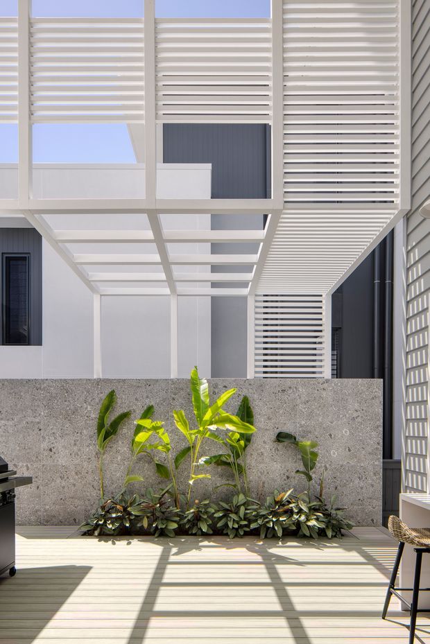 A bright contemporary courtyard or terrace enclosed by a modern multi-storey building. White horizontal louvres and pergola-like screens create strong linear shadows across a pale timber deck. A textured grey concrete planter wall supports tropical planting, with a partial outdoor dining setting visible at the edges. The architecture features clean white framing, dark grey cladding, and extensive horizontal sun-shading elements.