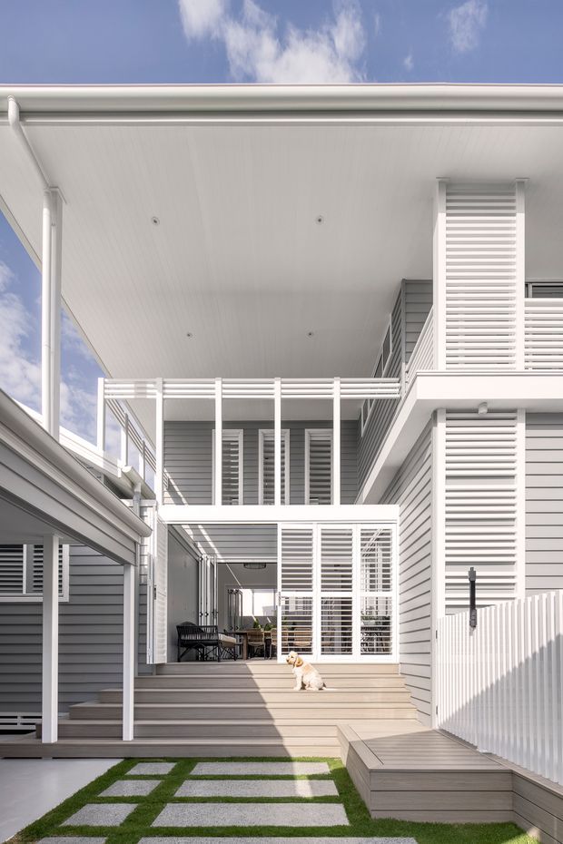 Bright exterior view of a multi-level residential home with white and pale grey horizontal weatherboard cladding, extensive white louvred screens, covered outdoor circulation spaces, timber decking and steps, and a geometric lawn-and-paver courtyard. The image looks upward through a narrow courtyard toward balconies and roof overhangs under a blue sky.