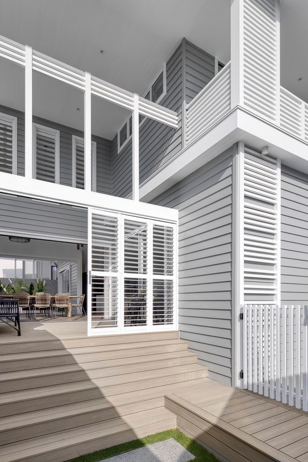 A bright multi-level residential exterior centered on a timber-look deck and broad steps. The facade is clad in horizontal grey weatherboards with extensive white louvred screens, balustrades, shutters, and framed glazing. An upper-level balcony and covered outdoor dining area are visible, creating a layered, breezy contemporary coastal composition.