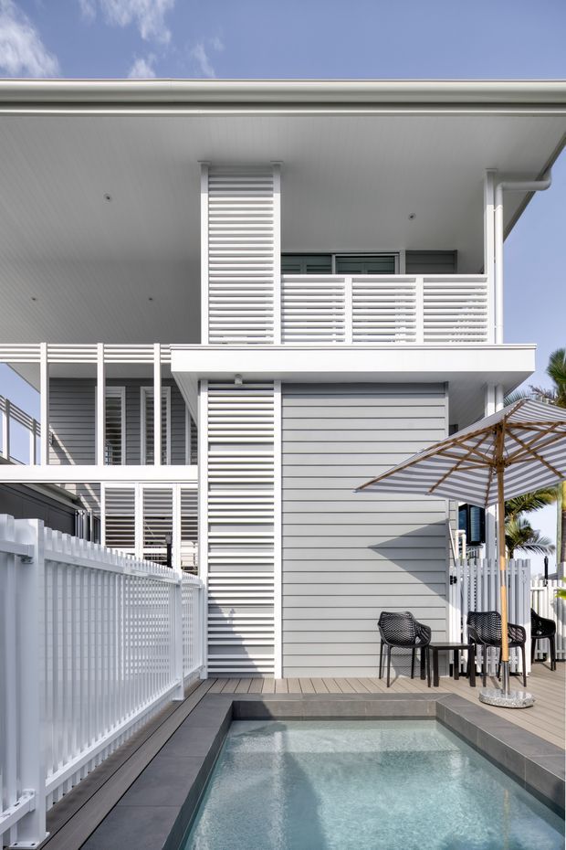 Bright exterior view of a modern multi-storey coastal-style residence with white horizontal cladding, extensive louvred screening, balconies, white fencing and a small swimming pool on a timber deck. Outdoor café seating and a striped umbrella sit beside the pool.