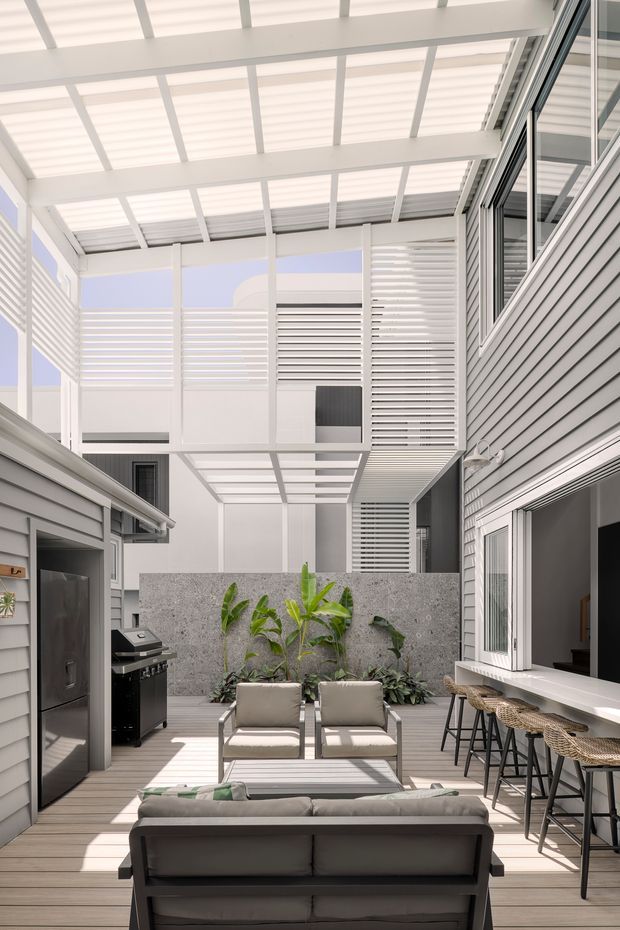 Bright contemporary enclosed courtyard entertaining area with a translucent covered pergola, white louvred privacy screens, grey weatherboard-style cladding, and extensive glazing. The space includes a timber-look deck, outdoor lounge seating, a built-in bar counter with stools, barbecue and refrigerator, plus a planted feature wall and concrete screening wall.