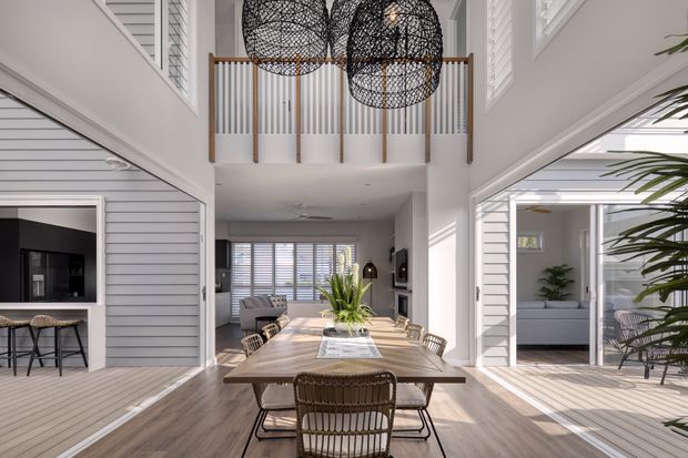Bright, spacious open-plan dining area with a large timber dining table, woven pendant lights, and a double-height ceiling. White horizontal wall panelling, extensive plantation shutters, and a timber-and-white balustrade create a refined coastal interior. A kitchen/bar nook and living areas are visible beyond the dining space, with strong daylight entering through large glazed openings.