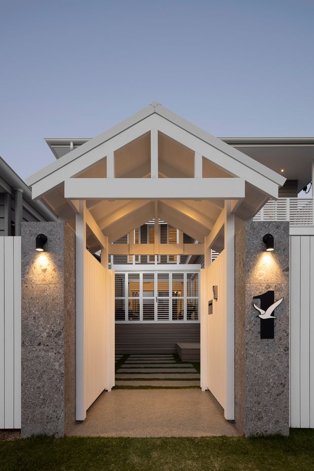 A symmetrical covered entrance passage leading into a residential property. The entry features a prominent white gabled timber structure with exposed rafters and warm timber lining, flanked by grey stone-clad pillars with black wall lights and decorative signage. Beyond the passage is a landscaped stepping-stone path and a glazed, louvred building entrance. The image is photographed from outside at dusk, with warm architectural lighting highlighting the sheltered walkway and pitched roof form.
