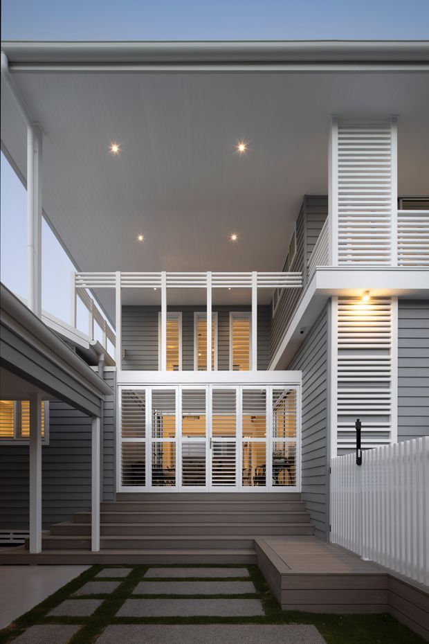 Exterior view of a compact multi-level contemporary residence centred on a sheltered courtyard and covered deck. The facade combines grey horizontal weatherboard cladding with extensive white-framed glazing, white horizontal louvres, balustrades and fencing. Recessed ceiling lights illuminate the covered outdoor areas, with timber decking and concrete stepping pavers visible in the foreground.