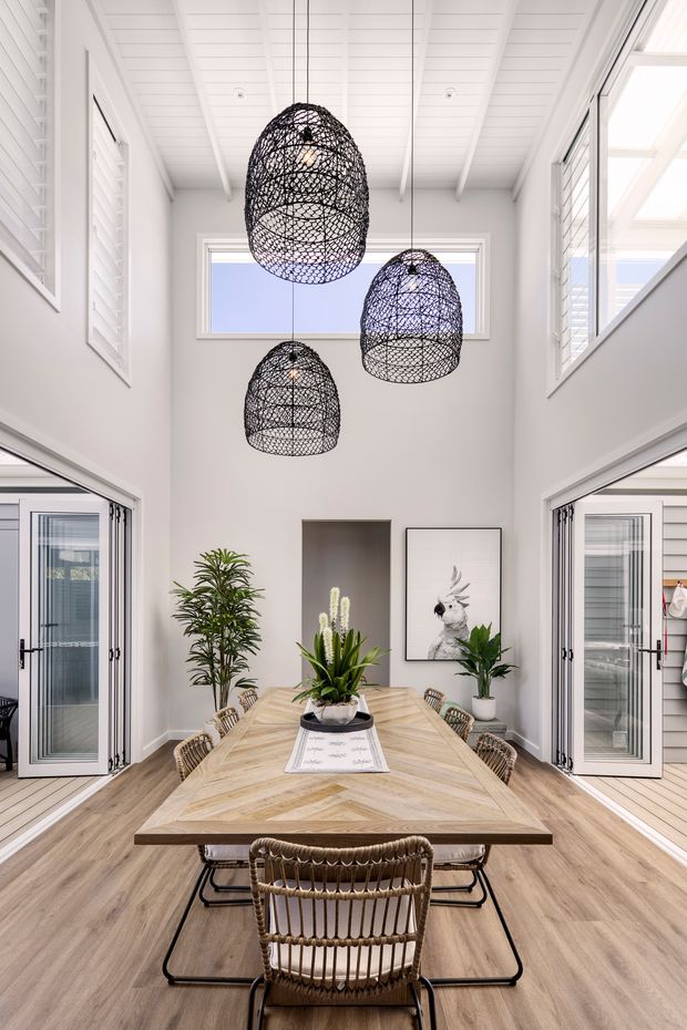 Bright dining room with a long light timber chevron-patterned dining table and woven chairs, arranged beneath three large black woven pendant lights. The room has white walls, a white exposed-beam pitched ceiling, high clerestory windows, louvre shutters, wide glazed openings, indoor plants, and light timber flooring. A large artwork and planted niches add visual interest. The space feels airy, warm, and coastal in character.