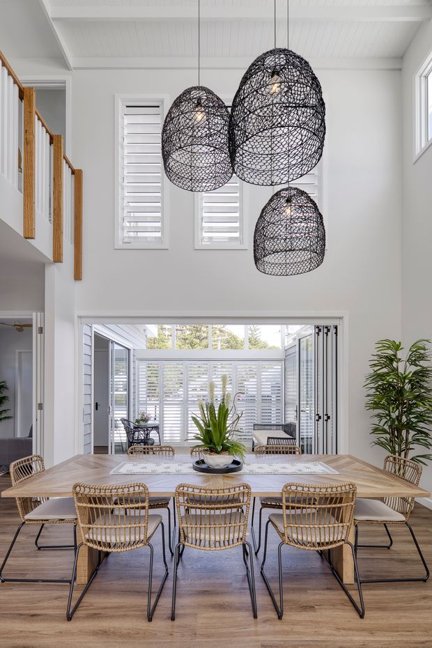 Bright double-height dining area with a large timber dining table surrounded by woven rattan dining chairs. Three oversized black woven pendant lights hang from a white paneled ceiling. White plantation shutters, extensive glazing, and an open connection to a leafy enclosed outdoor area create an airy, contemporary coastal feel. A timber-and-white stair balustrade is visible at left, with potted greenery adding natural texture.