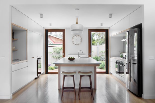 Bright, compact contemporary kitchen with a central island and two timber stools. Full-height white cabinetry lines both sides, incorporating a recessed open-shelf display niche on the left and integrated stainless-steel appliances on the right. The island has a pale smooth benchtop with an undermount sink and chrome tap. Timber-framed glazed doors at the rear open to a small garden, while a simple white pendant light, recessed ceiling lights, and a round wall clock complete the clean, functional interior.