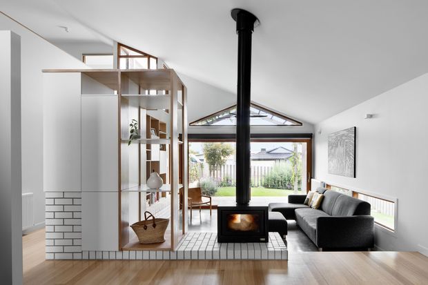 Bright contemporary living room in a renovated or extended home, featuring a steeply pitched ceiling, exposed timber-framed glazed gable, large rear glazing, polished timber flooring, a black freestanding wood-burning stove, and a built-in tiled hearth. A grey sofa and armchair face the stove, while an open timber-and-white shelving partition separates the living area from the adjacent space.