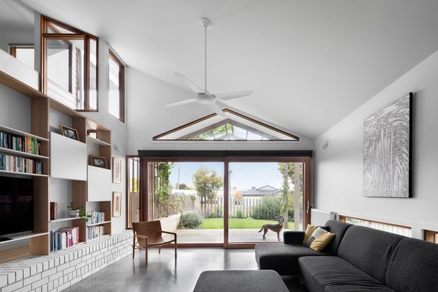 Bright contemporary living room with a high pitched ceiling and exposed triangular clerestory window above large timber-framed glazed doors opening to a garden. Built-in shelving and cabinetry line the left wall around a television, while a white brick feature wall, dark sectional sofa, timber armchair, polished concrete floor, and ceiling fan define the space.