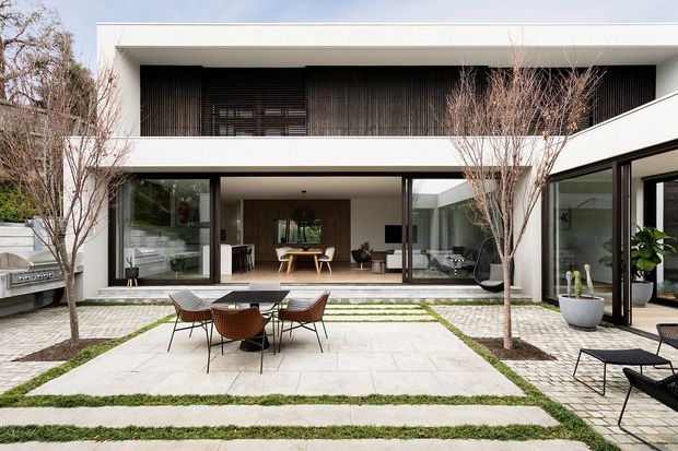 Contemporary two-storey residence viewed from a landscaped courtyard. The house has a crisp white rectilinear facade, broad floor-to-ceiling glazing, dark vertical timber screening on the upper level, and a sheltered ground-floor living area opening onto a paved outdoor dining terrace. Linear grass strips, pale stone paving, mature trees, outdoor furniture, and a built-in barbecue contribute to a refined indoor-outdoor setting.