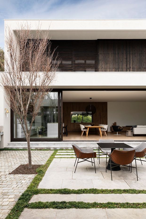 A refined contemporary two-storey house viewed from its landscaped rear courtyard. The white rectangular facade has a deep upper-level opening screened with dark vertical and horizontal timber slats, while large glazed sliding doors open the ground-floor living and dining area onto a paved outdoor terrace. The interior includes timber wall panelling, a dining table, pendant light, sofa and lounge seating. The courtyard features pale stone paving, bands of grass, cobblestone paths, a slender deciduous tree and dark woven outdoor furniture.
