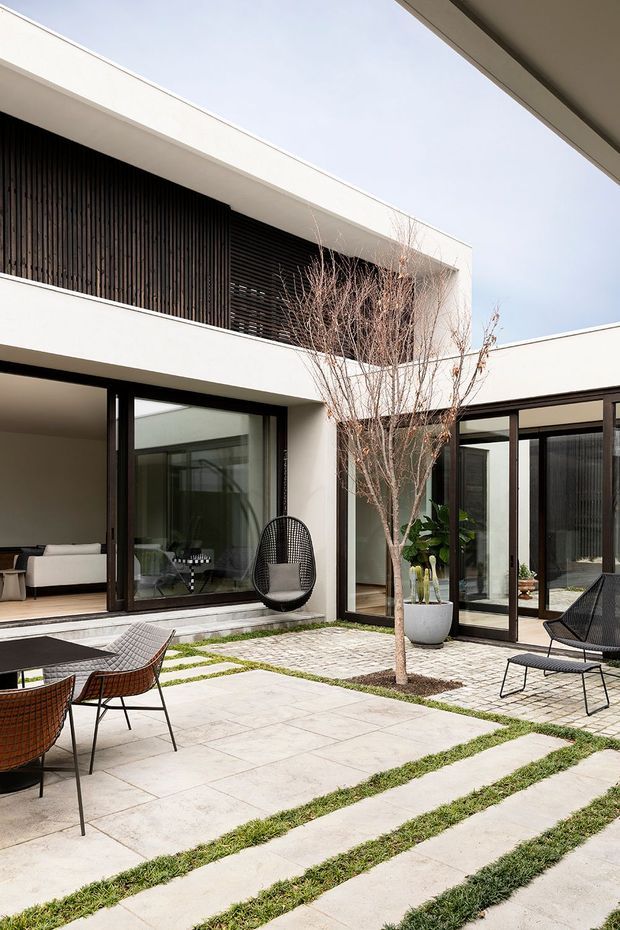Modern multi-level courtyard house with crisp white rendered walls, dark timber vertical screening, and large dark-framed glazed openings surrounding a paved outdoor courtyard. The courtyard features large pale stone or concrete pavers separated by narrow strips of grass, a small deciduous tree, planters, and black woven outdoor lounge and dining furniture. The composition emphasizes the private central courtyard and strong indoor-outdoor connection.