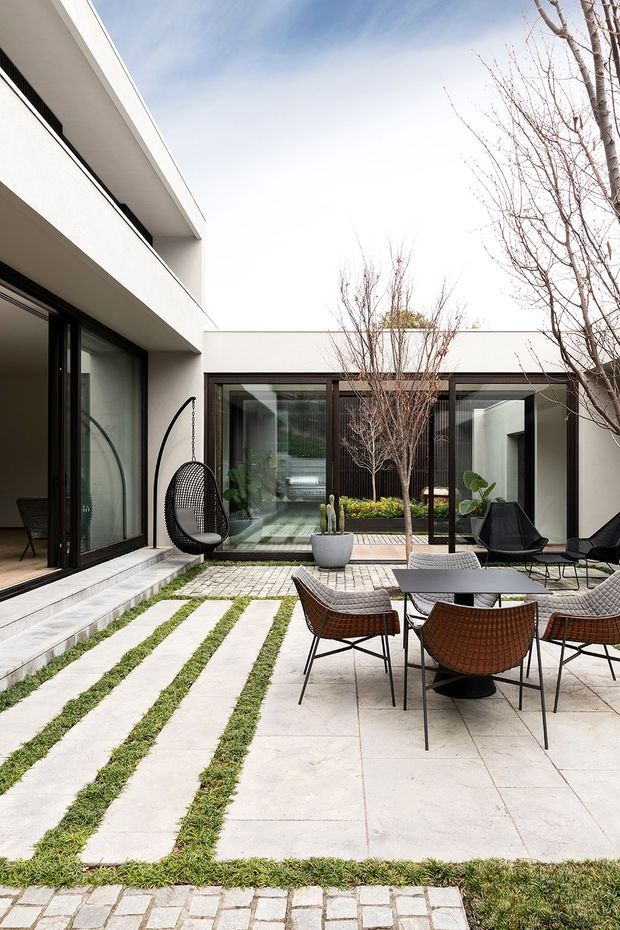 A contemporary multi-level courtyard residence viewed from an outdoor paved terrace. White rendered walls and deep projecting balconies frame dark-framed floor-to-ceiling glazing. The courtyard uses large pale concrete or stone pavers separated by narrow strips of grass, with a smaller cobbled border, mature bare-branched trees, potted plants, and a dining setting of woven outdoor chairs and a low table. A hanging egg chair sits beside the open living area, while glazed doors reveal the interior and a planted courtyard beyond.